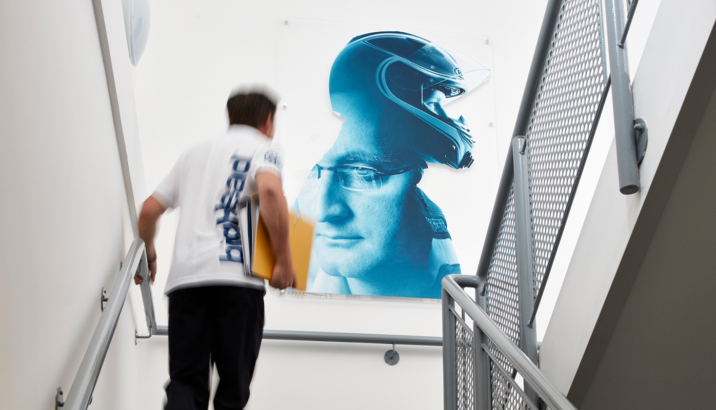 Image shows a large brand image on a staircase wall. The branded image is a montage of two heads that portray the process from high-performance design to high-performance motorsport.