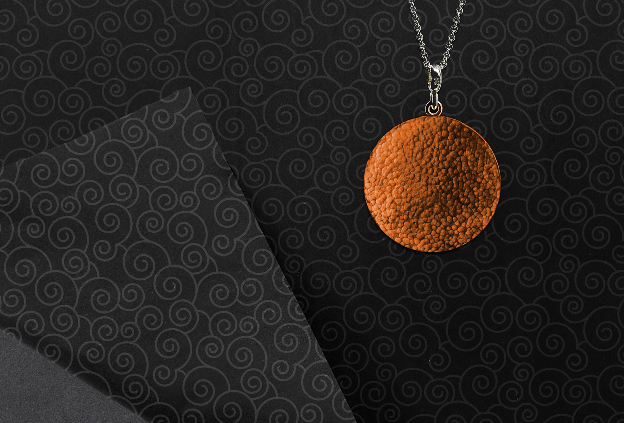 Close-up of a bronze pendant necklace sat on dark coloured branded tissue paper depicting illustrative patterns