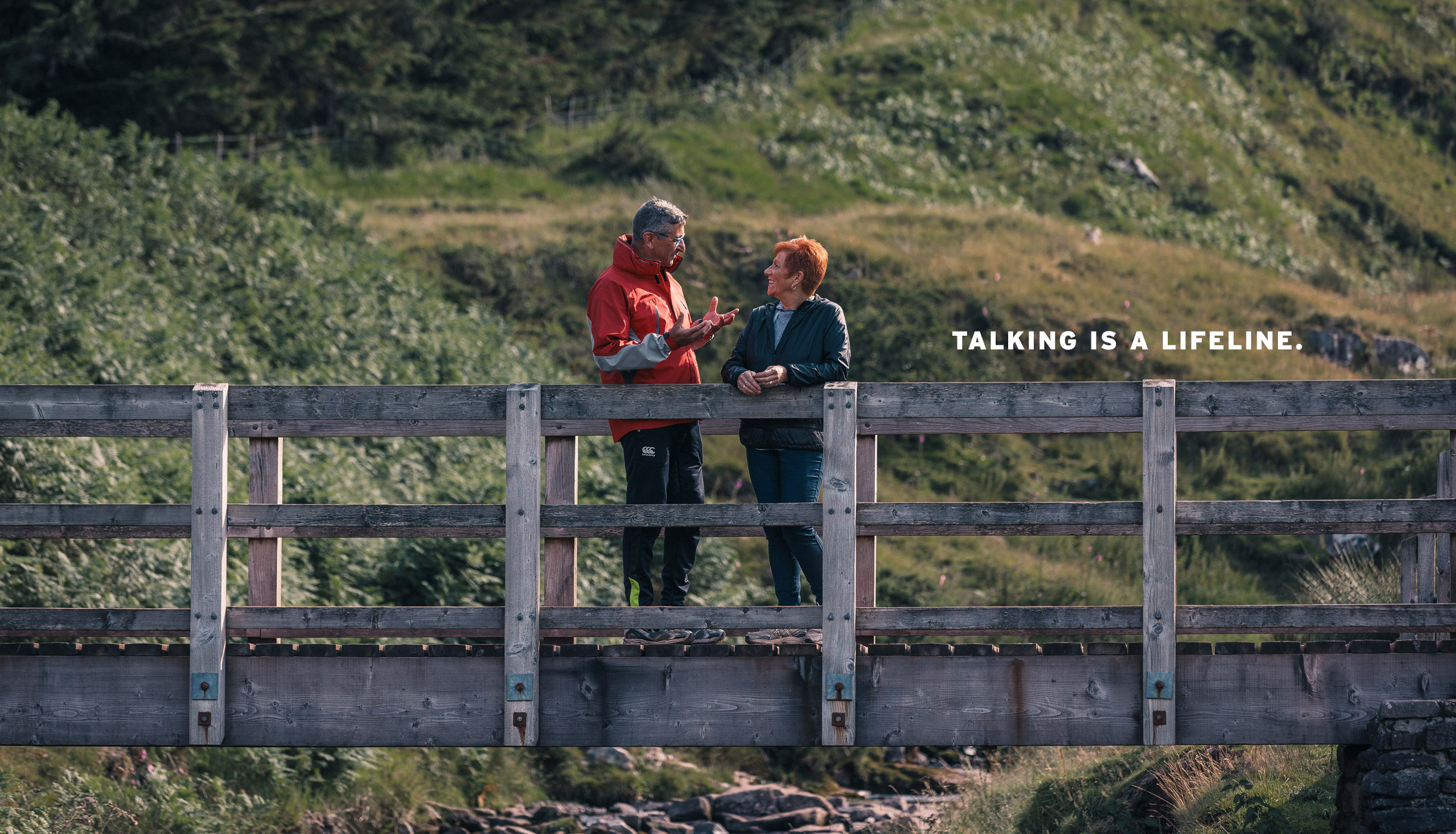 Man and woman in discussion, stood on a wooden bridge amongst mountains with the message 'Talking is a lifeline'