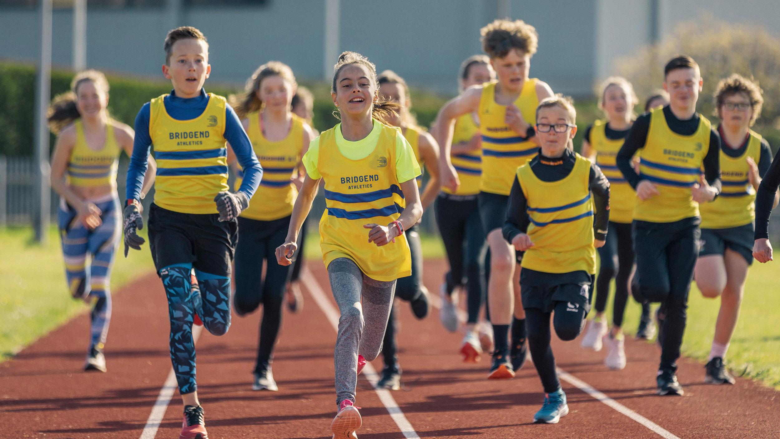 Photo of junior athletes wearing branded athletic club vests sprinting on running track